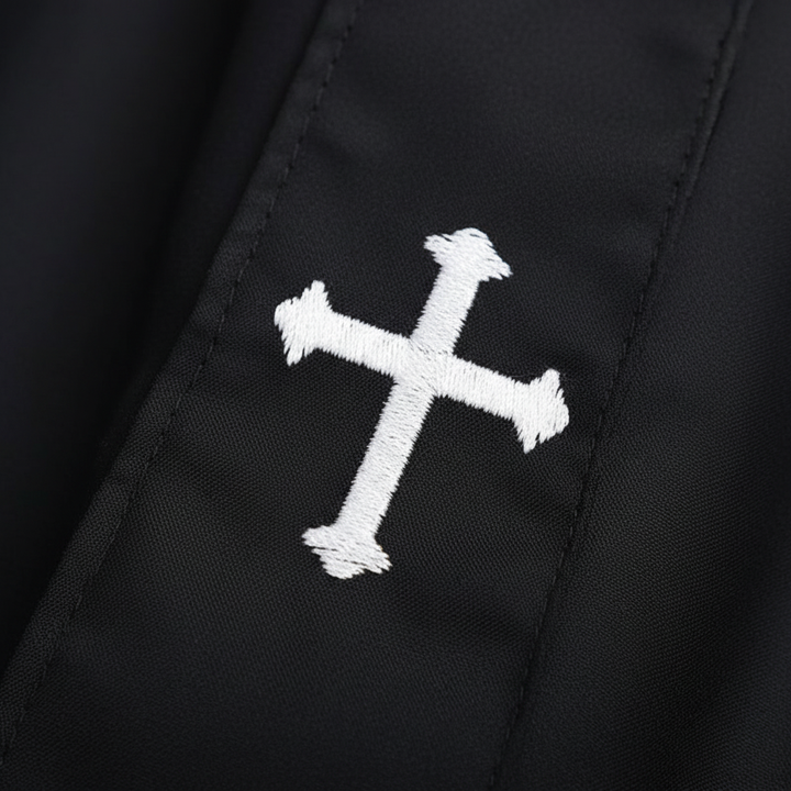 Close-up detail of cassock robe lower hem showing white cross embroidery on black fabric, white background
