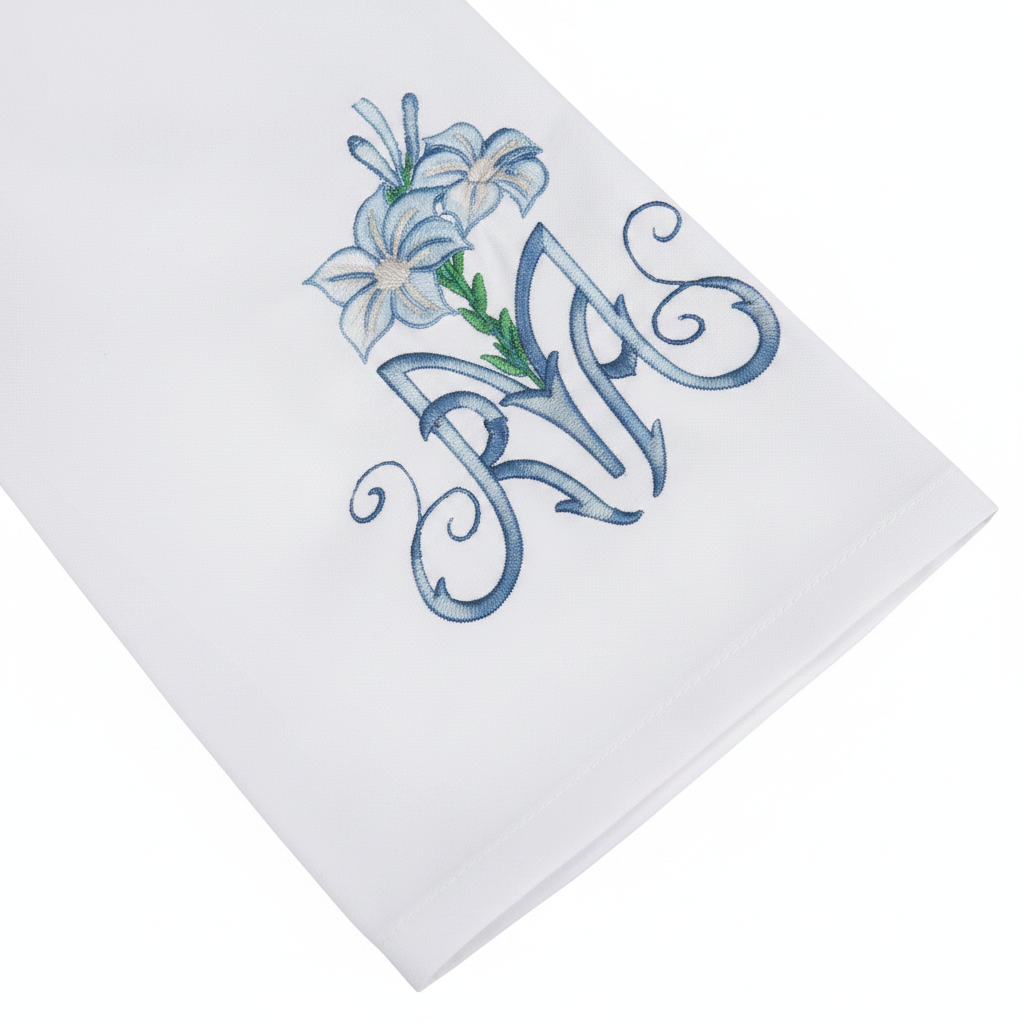 Close-up detail of white alb robe sleeve cuff showing blue lily and floral scroll embroidery, white background