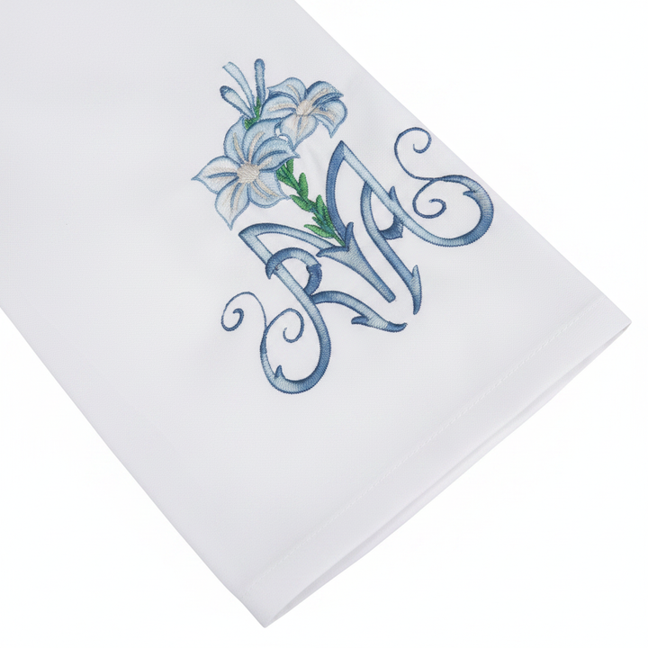 Close-up detail of white alb robe sleeve cuff showing blue lily and floral scroll embroidery, white background