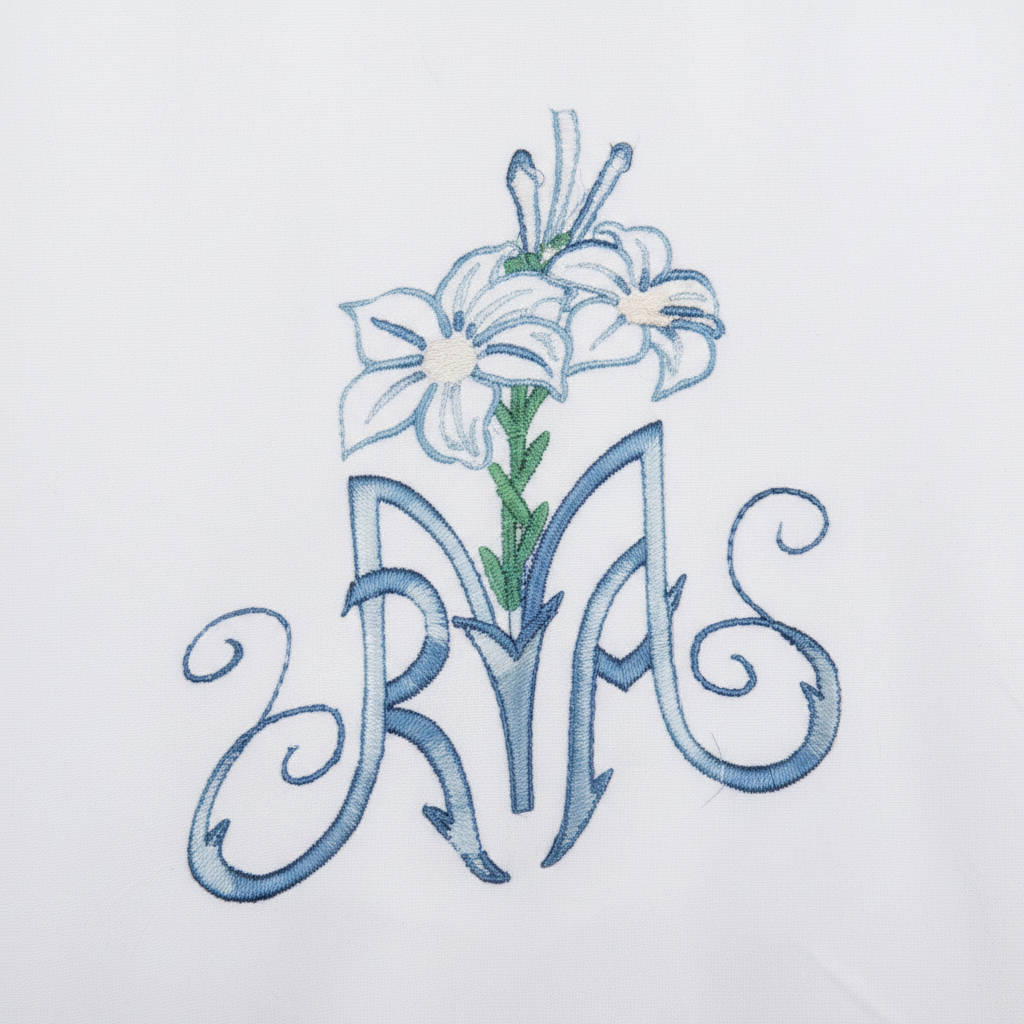 Close-up detail of white alb robe lower front hem showing blue AM monogram and lily embroidery, white background